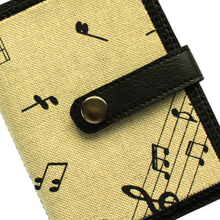 Load image into Gallery viewer, Close up of a notebook with musical notes fabric on a beige background stitched on to black faux leather with tab closure and metal rivet fastener