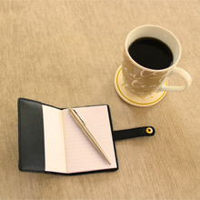 Load image into Gallery viewer, Black notebook with cover open with a pen on ruled paper next to a cup of coffee