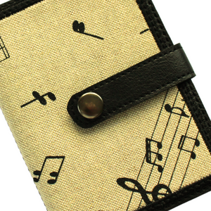 Close up of a notebook with musical notes fabric on a beige background stitched on to black faux leather with tab closure and metal rivet fastener