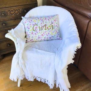 Cwtch cushion stitched in green on floral fabric on a chair with a white throw
