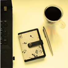 Load image into Gallery viewer, Notebook with musical fabric on a desk next to a laptop, pen and cup
