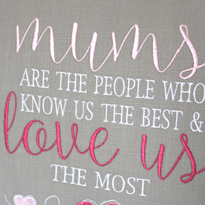 Close up view of the embroidered stitching of the Mums Love Us the most embroidered cushion text stitched in white, light pink and dark pink on grey fabric
